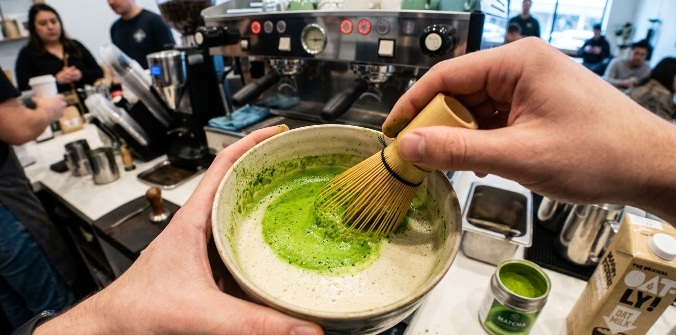 A vibrant green matcha powder being whisked into oat milk in a cafe setting.