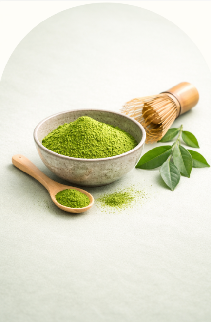 Vivid green matcha powder in a ceramic bowl with a bamboo whisk, wooden spoon, and fresh tea leaves on a soft sage background.