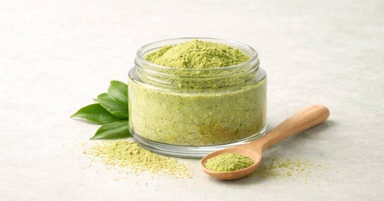 Glass jar of matcha scrub exfoliant with visible powder texture, wooden spoon, and fresh tea leaves on a pale background.
