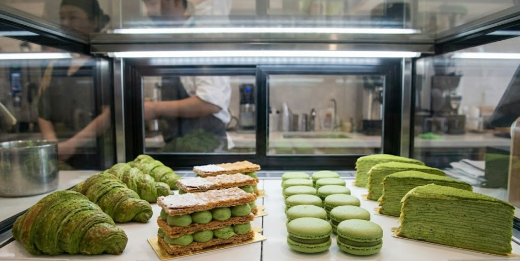 Close-up of matcha croissants, macarons, and crepe cake in bakery case.